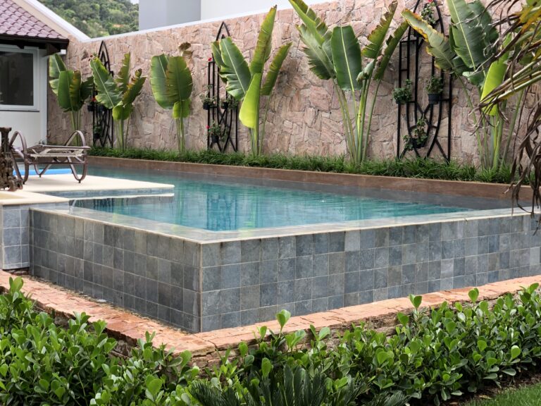 Piscina elevada com revestimento externo em pedra e paisagismo tropical, projeto da Ramiro Piscinas em Rio do Sul
