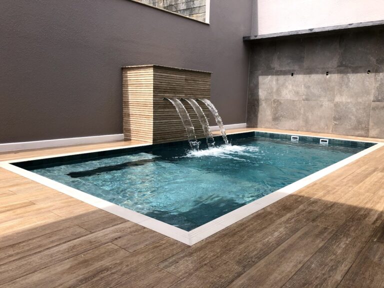 Piscina de alvenaria com parede de água em cascata e deck de madeira, projeto da Ramiro Piscinas
